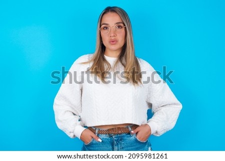 Portrait of lovely funny Young hispanic girl wearing white knitted sweater over blue background sending air kiss