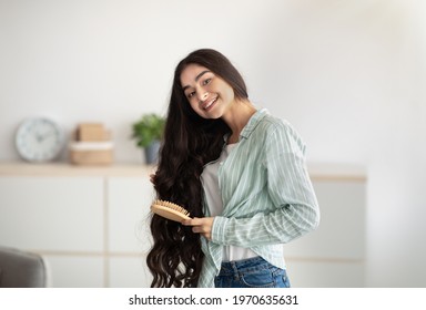 Portrait Of Lovely Indian Lady Brushing Her Wavy Long Hair, Using Wooden Brush Indoors. Cheerful Young Woman Taking Care Of Herself At Home. Domestic Spa Salon, Wellness And Beauty Concept