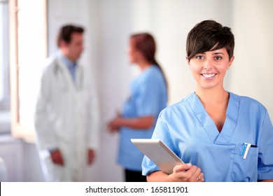 Portrait Of A Lovely Latin Nurse On Blue Uniform Standing And Smiling On Medical Team Background