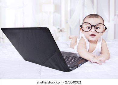 Portrait of lovely male baby lying on bedroom with laptop while wearing a round glasses and looking at camera