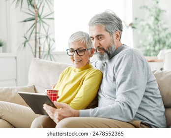 Portrait of a lovely senior mature couple together holding a tablet on sofa at home - Powered by Shutterstock