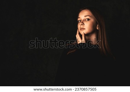 Portrait of lovely teenager cover girl posing in black at dark background, pensive looking at camera. Teen girl with dimples 12-13 year old isolated in shadow. Generation A concept. Copy ad text space
