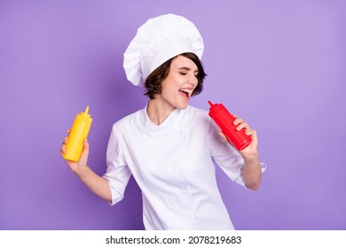 Retrato de un agradable chef de moda y alegre cantando botellas de ketchup divertiéndose en un café rápido aislado sobre fondo violeta violeta de color morado brillante