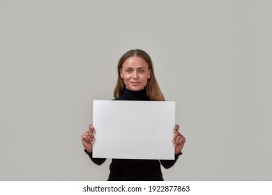 Portrait of lovely young caucasian woman smiling at camera, holding white blank banner in front of her while standing isolated over gray background. Advertisement concept. Copy space