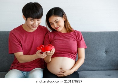 Portrait of a lovely young family couple. A man and a pregnant woman wearing a red T-shirt sitting on a couch smiling and looking at each other holding a red baby shoe.