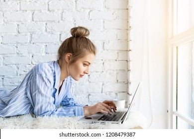 Portrait Of Lovely Young Female Using Trendy Laptop For Work From Home. Sweet Blonde Model In Pyjamas With Tied Up Hair. Technology And Lifestyle Concept. Copy Space In Right Side