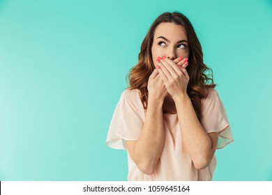 Portrait of a lovely young girl in dress standing with mouth covered and looking away at copy space isolated over blue background