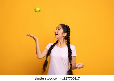 Portrait of lovely young indian woman playing with ripe green apple, juggling and throwing fruit up in the air standing isolated on yellow studio background. Beautiful lady promoting healthy nutrition