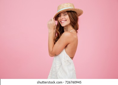 Portrait Of A Lovely Young Lady In Summer Dress And Hat Posing While Standing And Laughing Isolated Over Pink Background