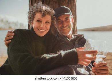 Portrait Of Loving Mature Couple Sitting Together With A Glass Of Wine. Man And Woman Camping Near A Lake On A Summer Day.