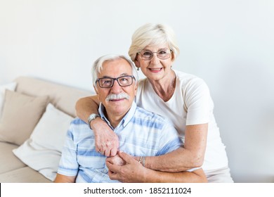 Portrait Loving Older Wife Hugging Husband Sitting On Cozy Couch. Happy Senior Mature Couple Smilling And Looking At Camera, Posing For Family Photo At Home. Elderly Couple Feeling Happy