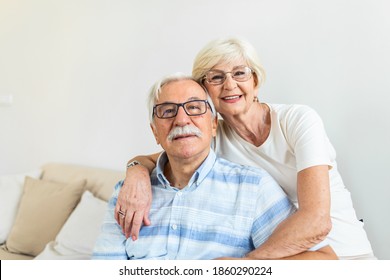 Portrait Loving Older Wife Hugging Husband Sitting On Cozy Couch. Happy Senior Mature Couple Smilling And Looking At Camera, Posing For Family Photo At Home. Elderly Couple Feeling Happy