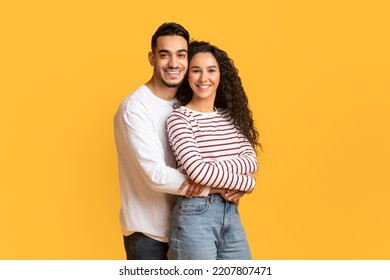 Portrait Of Loving Young Arab Man And Woman Embracing And Smiling At Camera, Romantic Middle Eastern Couple Hugging While Posing Together Over Yellow Background In Studio, Copy Space