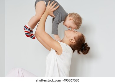 Portrait of loving young mother playing with her kid. Playful woman holding little cute son above her head. Calm caring mommy in white top touching forehead of baby with her nose, making him smile.