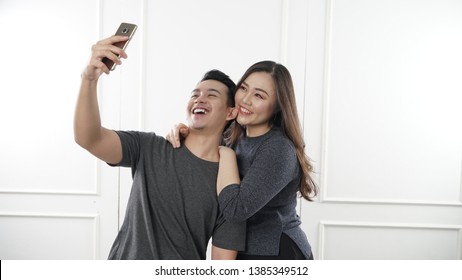 Portrait Of A Loving Young South East Asian Couple Hugging While Standing Together And Taking A Selfie Isolated Over White Background