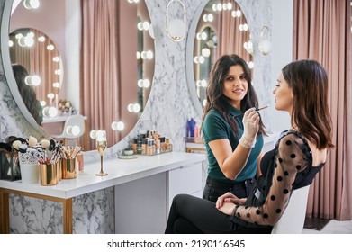 Portrait Of Makeup Artist Smiling To Camera While Doing Professional Makeup In Visage Studio. Woman Sitting At Dressing Table While Female Beauty Specialist Applying Lipstick With Cosmetic Brush.
