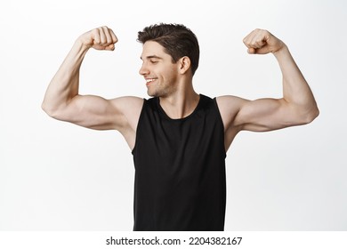 Portrait of male athlete, smiling sportsman flexing biceps, looking pleased at muscle biceps, workout in gym, standing over white background.