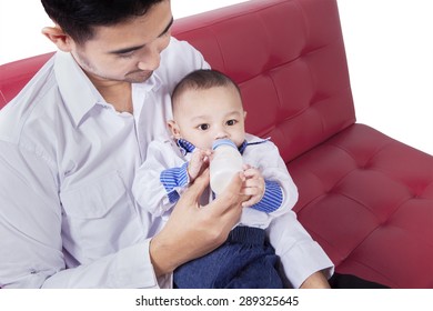 Portrait of male baby with a cute face, drinking milk from bottle with his dad on the sofa