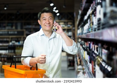 Portrait Of Male Buyer In Supermarket, Asian Smiling And Looking At Camera Talking Cheerfully On Phone, Choosing Wine In Liquor Department