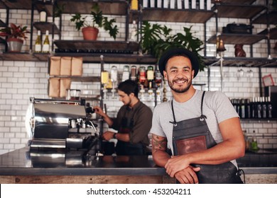 Portrait Of Male Coffee Shop Owner Standing At The Counter With Barista Working In Background Making Drinks.