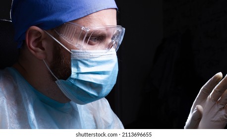 Portrait Of A Male Doctor In A Mask, Glasses And Uniform On A Dark Background. Medic Works In The Office.