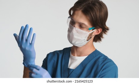 Portrait of male doctor wearing medical mask and gloves preparing for a working day in hospital. Young intern dressed in uniform and protected glasses standing over white background