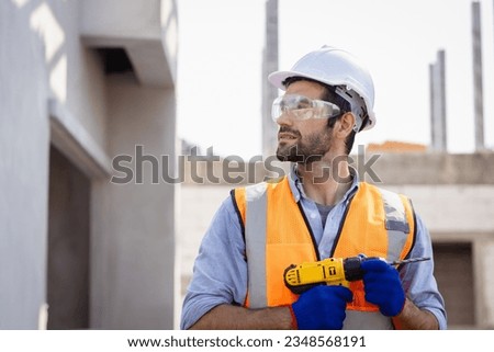 Portrait  male engineer builder renovating with drill in mask helmet and glove.Handsome Technician using cordless drill in safety first concept.House building under construction site.