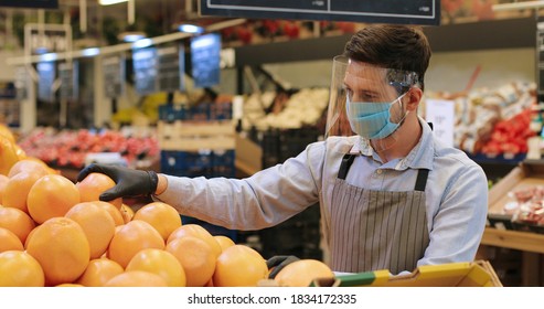 Portrait Of Male Food Store Employee In Face Protection Shield And Apron Standing In Store And Sorting Fruits. Handsome Caucasian Man Seller In Mask Working Indoor. Supermarket Concept