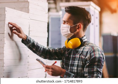 Portrait Male Foreman Inspector In Medical Face Mask And Protective Headphones Checking Woodwork Stock At Factory Storage. Man Supervisor Counting Wood Inventory. Warehouse Worker. COVID-19 Quarantine