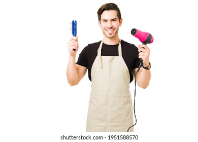 Portrait Of A Male Hair Stylist Working At A Beauty Salon. Professional Young Man Smiling While Making Eye Contact And Holding A Blow Dryer And A Brush