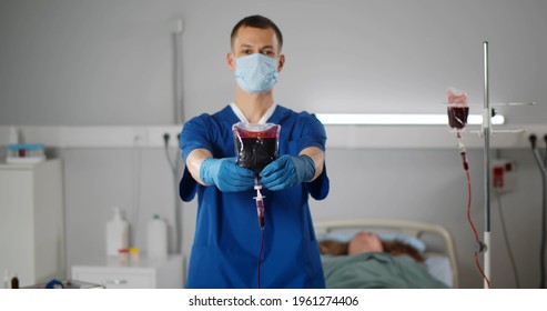 Portrait Of Male Nurse In Safety Mask Holding Blood Bag And Looking At Camera Doing Blood Transfusion To Patient In Hospital Ward. Healthcare And Medicine Concept