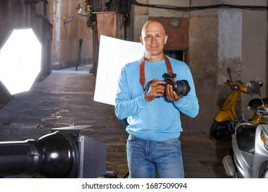 Portrait Of Male Photographer Standing With Camera Among Professional Photo Equipment On Old City Street 