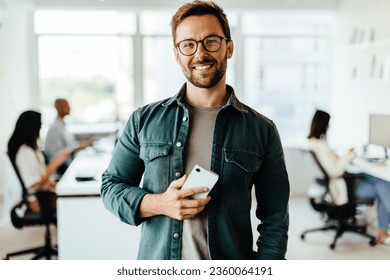 Portrait of a male professional standing in an office with his colleagues in the background. Business man looking at the camera while holding a mobile phone. - Powered by Shutterstock