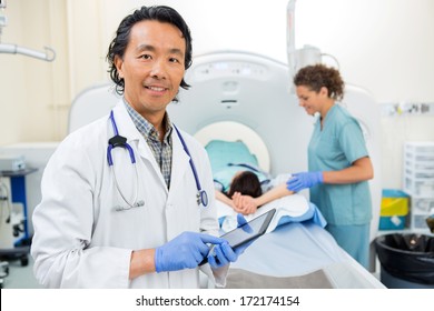 Portrait Of Male Radiologist Using Digital Tablet While Nurse Preparing Patient For CT Scan In Hospital