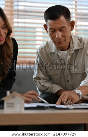 A portrait of a male senior client sitting on a couch in a house pointing out the agreement condition, for real estate, business, finance and insurance concept.