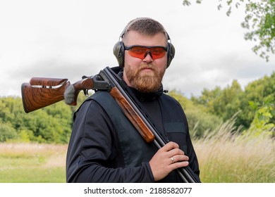 Portrait Of A Male Shooter 30-35 Years Old With A Sports Rifle On His Shoulder, Wearing Glasses And Tactical Headphones For Shooting Against The Background Of Nature.