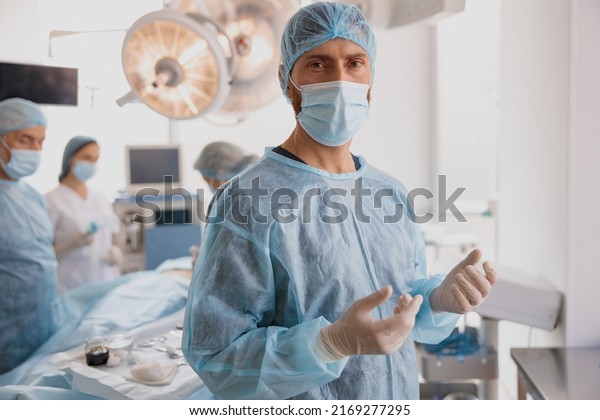 Portrait of male surgeon in mask\
and gloves standing in operating room, ready to work on\
patient