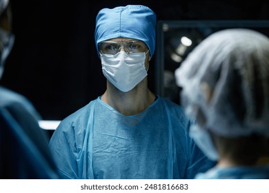 Portrait of male surgeon wearing scrubs and protective gear with medical team in operating room lit by blue light copy space - Powered by Shutterstock