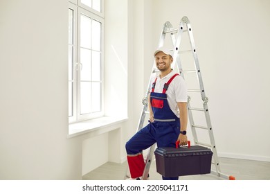 Portrait Of Male Worker Inside House. Happy Man In Work Uniform Overalls Standing In New Empty Room With White Walls, Holding Toolbox, Looking At Camera And Smiling. Repairs And Renovations Concept