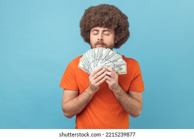 Portrait Of Man With Afro Hairstyle Wearing Orange T-shirt Smelling Dollar Banknotes With Pleasure Expression, Enjoying Big Money Profit And Rich Life. Indoor Studio Shot Isolated On Blue Background.