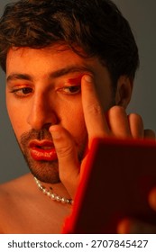 Portrait of a man applying makeup while looking into a small mirror, captured with warm studio lighting and a neutral background.