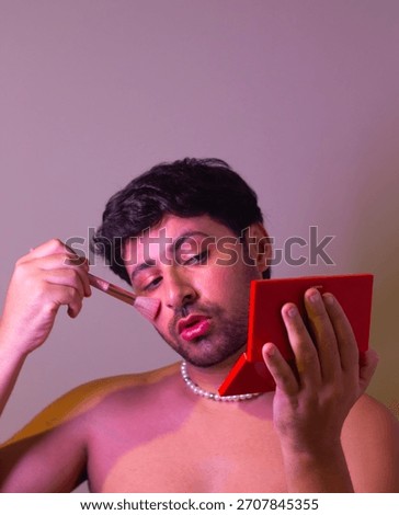 Portrait of a man applying makeup while looking into a small mirror, captured with warm studio lighting and a neutral background.