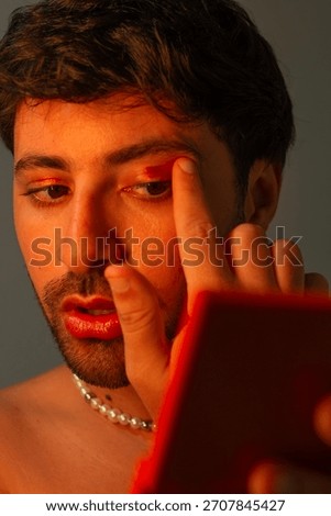 Portrait of a man applying makeup while looking into a small mirror, captured with warm studio lighting and a neutral background.