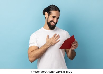 Portrait Of Man With Beard Wearing T-shirt Reading Letter From Red Envelope, Gets Greeting Card, Having Positive Romantic Expression, Being Pleased, Indoor Studio Shot Isolated On Blue Background.