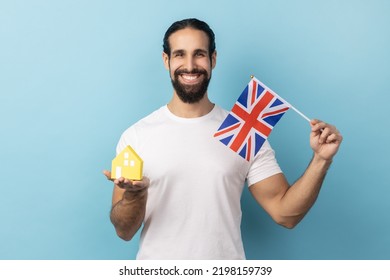 Portrait Of Man With Beard Wearing White T-shirt Holding British Flag And Paper House, Dreaming To Buy Accommodation In United Kingdom. Indoor Studio Shot Isolated On Blue Background.