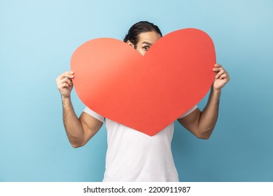 Portrait Of Man With Beard Wearing White T-shirt Peeking Out Of Big Read Heart With Flirting Look And Expressing Love, Greeting On Valentine's Day. Indoor Studio Shot Isolated On Blue Background.