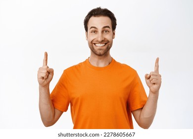 Portrait of man with beard and white smile, points fingers up, shows promo, logo upwards, stands in orange t-shirt.