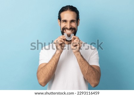 Portrait of man with beard in white T-shirt using teeth whitening braces, dental aligner retainer, dental clinic for beautiful teeth, treatment course. Indoor studio shot isolated on blue background.
