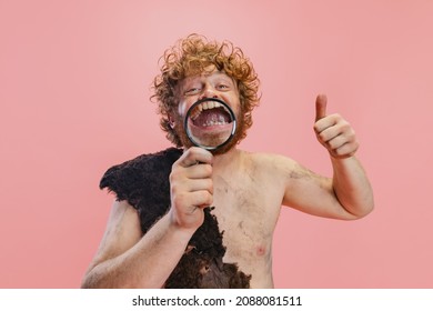 Portrait Of Man In Character Of Primitive Person, Neanderthal Holding Magnifier Enlarging Teeth Isolated Over Pink Background. Concept Of History, Modern And Ancient Combination, Evolution And Ad