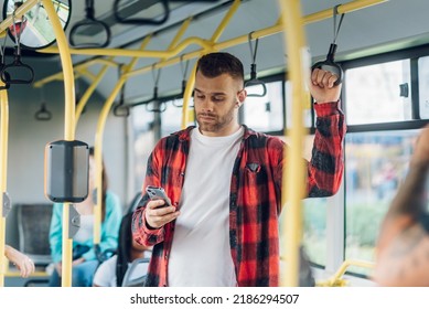 Portrait of a man in checkered shirt standing in a bus and using a smartphone. Everyday life and commuting to work by public transportation. Man is wearing airpods browsing social media - Powered by Shutterstock